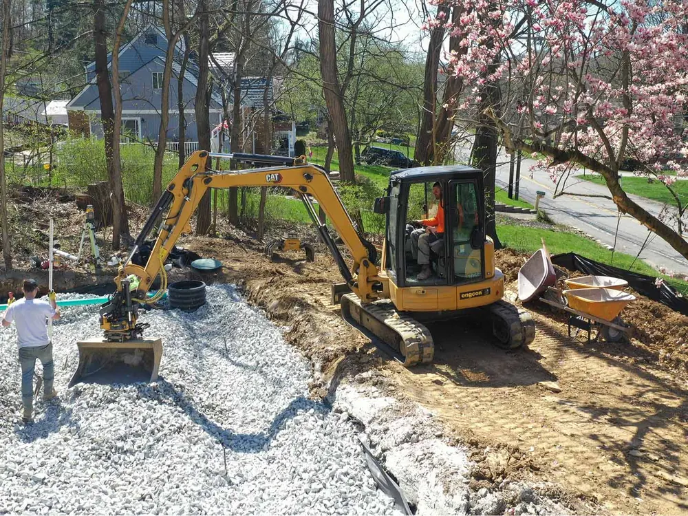 Mini excavator spreading drain stone over a yard drainage bed beside a house, french drain installation