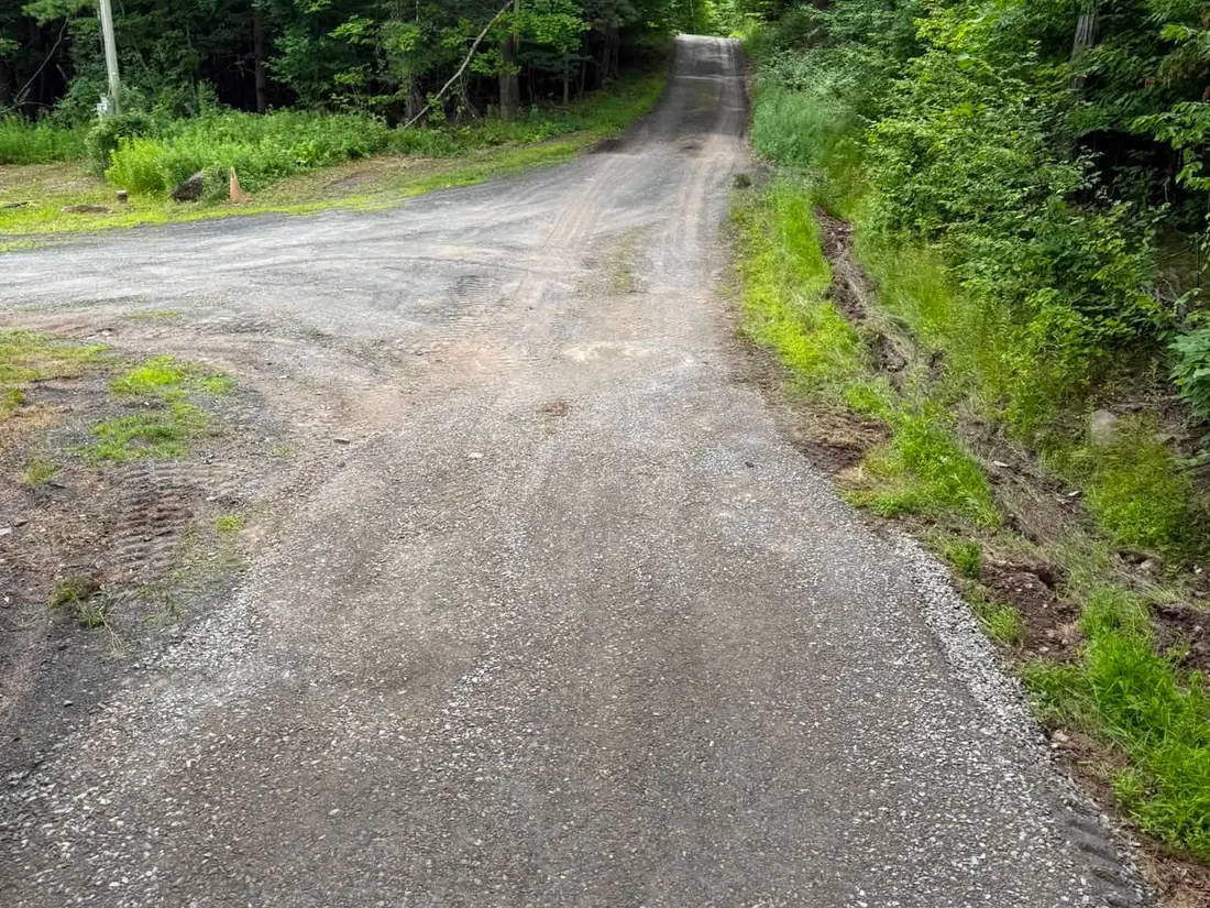 Gravel driveway with a roadside ditch cut along its edge to carry runoff, drainage work