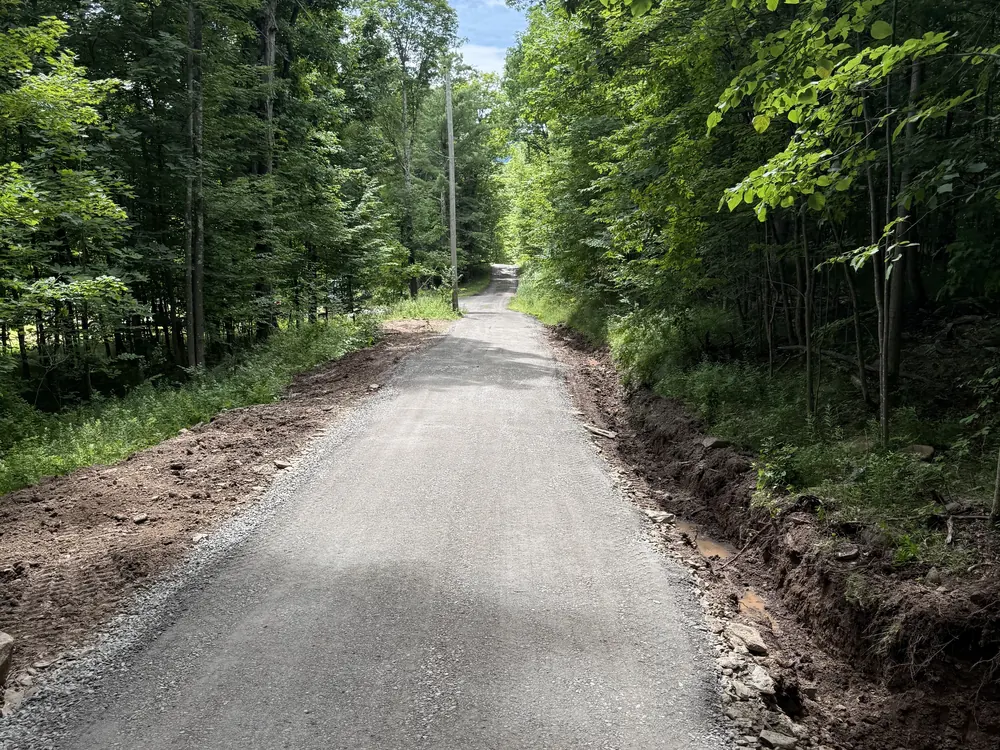 Fresh drainage ditch dug along a gravel driveway, water standing in the ditch, French drains and drainage work