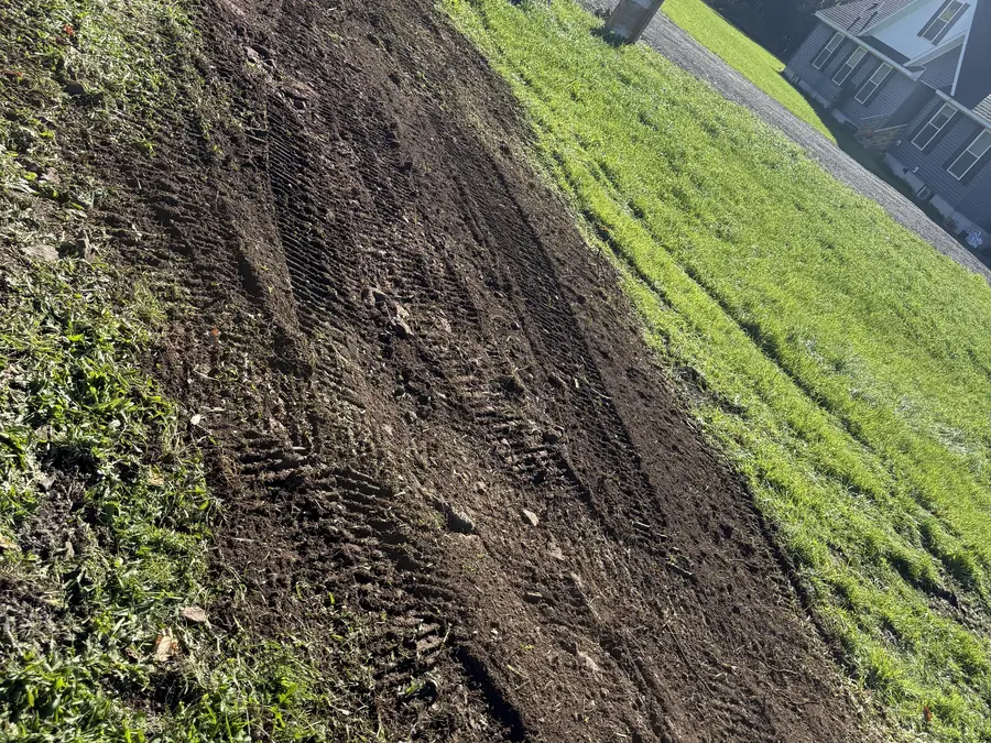Regraded strip of topsoil with track marks across a sloped lawn beside a house, land grading