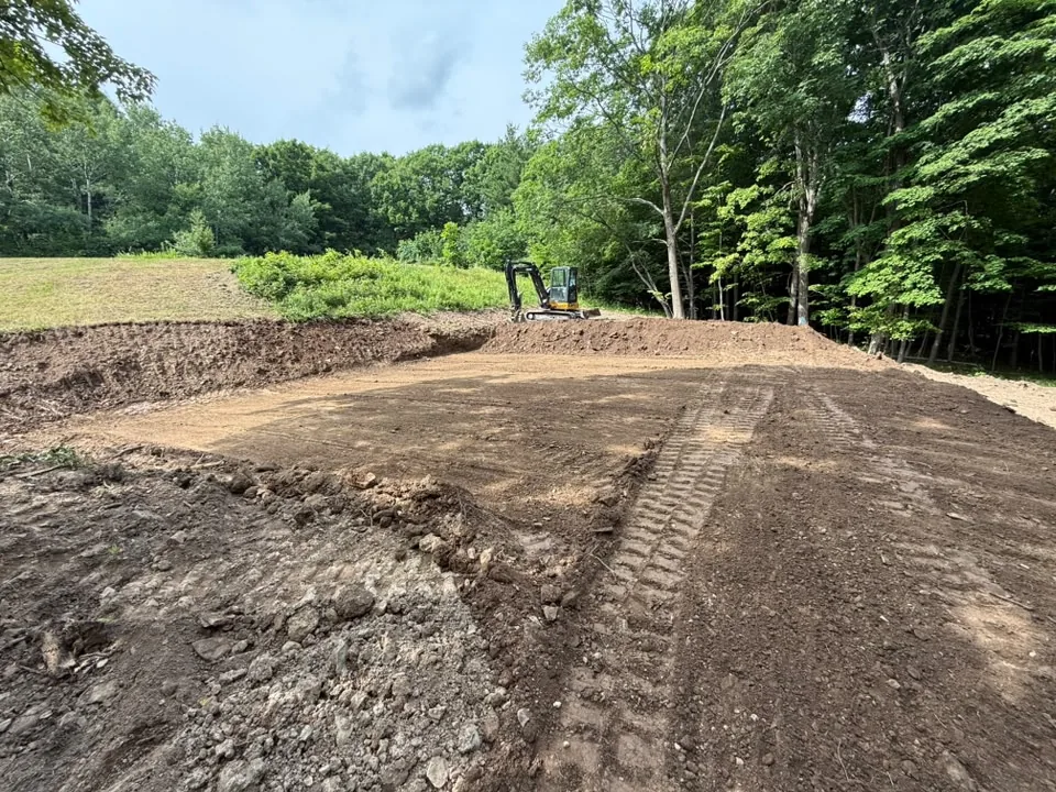 Site preparation with a mini excavator cutting a level pad into a hillside at the edge of a field