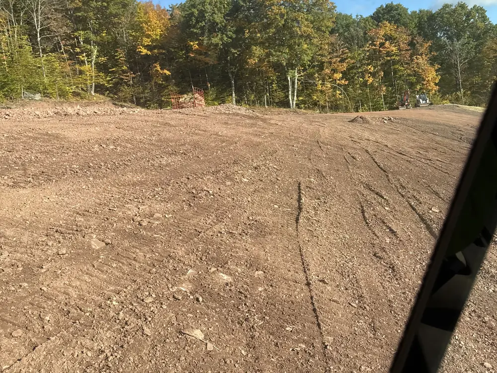 Graded pad of red shale soil seen from the machine cab, an excavator and loader at the far edge